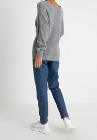 Personne portant un pull gris côtelé, un jean bleu et des baskets blanches, se tenant devant un fond blanc uni.