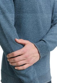 Blau strukturiertes Langarmhemd mit enger Passform, ausgestattet mit einem gerippten Bündchen und nahtlosem Design, präsentiert mit einer Hand, die auf dem Arm ruht.