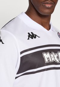 Hvid sportstrøje med krave, sorte stribede detaljer og Kappa-logo på skuldrene. Har tekstureret stof og fed "MAK"-tekst.