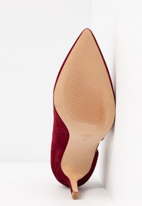Chaussure à talon haut en daim rouge à bout pointu vue par la semelle, reposant verticalement contre une surface blanche.