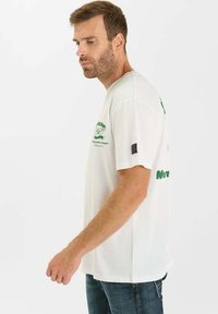 Witte katoenen t-shirt met groene grafische tekst en ontwerp. Korte mouwen, ronde hals en zwart label op de linkermouw.