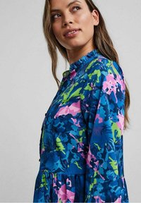 Šaty s květinovým vzorem v modré, zelené a růžové barvě. Mají řasený límec a knoflíkové detaily, s přiléhavým živůtkem a rozšířenou sukni.