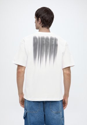 Homme aux cheveux courts et foncés portant un t-shirt blanc avec un texte noir flou "DIESEL" dans le dos et un jean bleu, de dos.