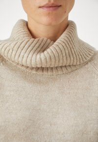 Maglione beige a maglia con colletto alto spesso e righe a costine. Texture morbida e vestibilità ampia, caratterizzato da un delicato motivo melangiato su tutta la superficie.