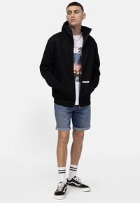 Črna jopa s kapuco z zadrgo, priložnostna majica s potiskom, denim kratke hlače in črni supergi z belimi črtami na nogavicah. Preprost, sproščen kroj.