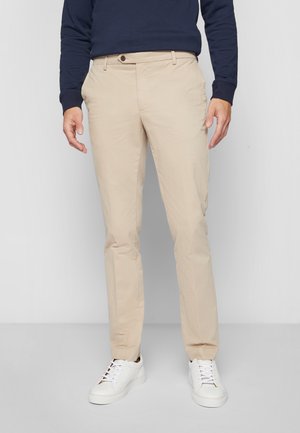 Mann trägt beigefarbene Slim-Fit-Chinos, weiße Sneakers und ein navyblaues Langarmshirt, steht vor einem schlichten grauen Hintergrund.