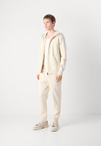 Jonge man staat, gekleed in een beige hoodie, bijpassende joggers, een licht tanktop en beige sneakers, tegen een eenvoudige witte achtergrond.