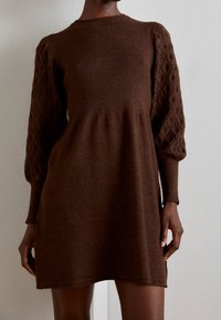 Vestito a maglia marrone con corsetto a coste e maniche a palloncino in maglia a treccia. Texture morbida, forma leggermente svasata, lunghezza a metà coscia.