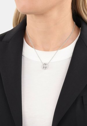 Collana in argento con pendente a forma ovale caratterizzato da un intricato design intrecciato, indossata sopra un top bianco e un blazer nero.