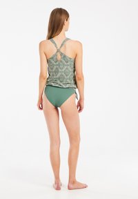 Maudymosi viršus su sudėtingu žaliu geometriniu raštu, su rakto skylutės nugaros detale, derinamas su vienspalviais žaliais bikini apatiniais. Sklandžios medžiagos tekstūra.