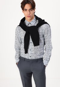 Lichtblauw button-up overhemd met bloemenpatroon, zwarte trui om de nek gedrapeerd, grijze broek; gladde stof, slim fit.