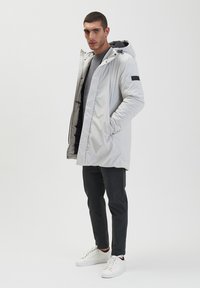 Lichtgrijze, gevoerde jas met een ritssluiting en capuchon, voorzien van zijzakken en een zwarte accent op de mouw. Gekocht met donkere broeken en witte sneakers.
