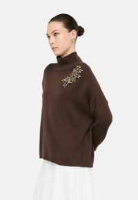 Brązowy sweter z dzianiny z wysokim kołnierzem, ozdobiony kwiatowym haftem w kolorze złotym i niebieskim na lewym ramieniu. Luźny fason z bocznymi żeberkami.