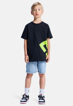 T-shirt en coton noir avec un grand motif vert néon, associé à un short en denim clair et des baskets noir et blanc. Texture plane et lisse.