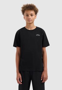 Tiener met krullend haar, gekleed in een eenvoudig zwart T-shirt met een klein wit logo op de linkerborst, staand tegen een lichtgrijze achtergrond.