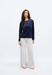 Γυναίκα με μακριά σγουρά μαλλιά που φοράει μπλε βελούδινη μπλούζα με μακριά μανίκια και φαρδιά μπεζ παντελόνια, όρθια, ξυπόλητη σε λευκό φόντο.