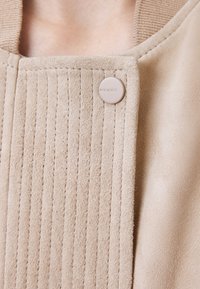 Giacca in suede beige con righe verticali e una texture liscia. Presenta un bottone rotondo beige con il nome del marchio.