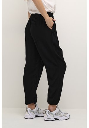 Persoon draagt zwarte joggers met elastische taille en witte sportschoenen, staat met één hand in de zak tegen een effen lichte achtergrond.