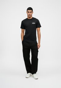 Schwarzes T-Shirt mit kurzen Ärmeln und einem weißen Logo auf der Brust, kombiniert mit schwarzen Cargo-Hosen und weißen Sneakers. Model steht vor einem neutralen Hintergrund.