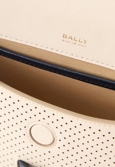 Κοντινό πλάνο στο εσωτερικό μιας κρεμ τσάντας από διάτρητο δέρμα που δείχνει το χρυσό λογότυπο Bally και το κούμπωμα με κουμπί.