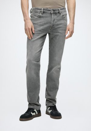 Homme portant un jean gris droit et des baskets noires avec des détails blancs et marron, debout devant un fond uni.