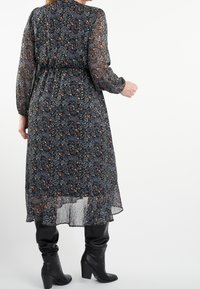 Robe maxi noire à motifs floraux, avec manches longues transparentes, taille cintrée et ourlet à volants. Associée à des bottes noires montantes à talons.