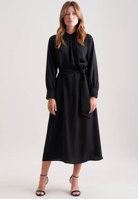 Donna che indossa un lungo vestito nero con un colletto alto, maniche lunghe, una cintura in vita e sandali neri con punta scoperta, in piedi contro uno sfondo semplice.