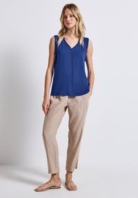 Blusa sin mangas azul con escote en V y detalle de costura frontal, combinada con pantalones tapered beige claros y sandalias planas con tiras.