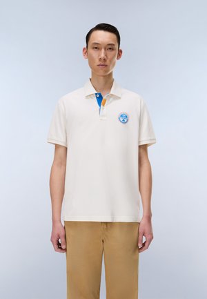 Giovane uomo che indossa una polo color crema con dettagli di bottoni blu e arancioni e pantaloni kaki, in piedi davanti a uno sfondo grigio chiaro.