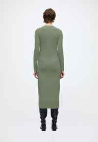 Alyvuogių žalios spalvos plisuota midi suknelė su ilgomis rankovėmis, apvalia iškirpte ir prigludusia silueto linija, derinama su juodais batais iki kelių.