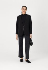 Svart blazer med en enkelt knapp, brukt over en svart topp. Kombinert med mørke jeans og svarte slip-on sko. Minimalistisk design, jevn tekstur.