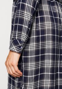 Una mano appoggiata accanto a una camicia a maniche lunghe a quadri blu navy e bianca, con polsini abbottonati.