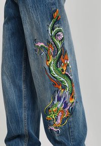 Blue denim jeans with a colorful embroidered dragon design featuring green scales and orange flames on the outer leg.