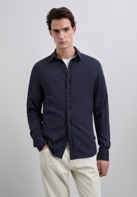 Navy button-up shirt with a soft texture, classic collar, and long sleeves, paired with light beige pants. Minimalist design, no visible patterns.