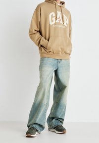 Sweat à capuche beige avec logo "GAP" blanc et accent rouge, associé à un jean large bleu clair. Complété par des baskets vertes et blanches.
