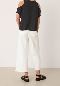 Personne portant un haut noir à épaules dénudées, un pantalon blanc taille large et court, et des sandales noires à boucle, se tenant sur un sol beige contre un mur blanc.