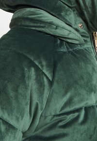 Grön sammetsägd puffjacka med en slät yta, kviltat mönster och dragkedja med en guldfinish.