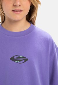 Camiseta de algodón morada con un logo gráfico central en negro y teal. Corte relajado con un cuello redondo clásico y una textura suave.