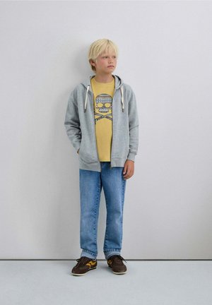 Sudadera gris con cremallera y cordones blancos, camiseta gráfica amarilla, jeans azules y deportivas marrones con acentos amarillos. Diseño y texturas simples.