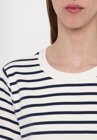 Femme portant une chemise blanche à rayures noires horizontales, montrant les lèvres et le cou sur un fond clair uni.