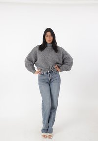 Grijze, oversized gebreide trui met een hoge halslijn, gecombineerd met lichtblauwe, high-waisted jeans. Model draagt lichtgekleurde sandalen.