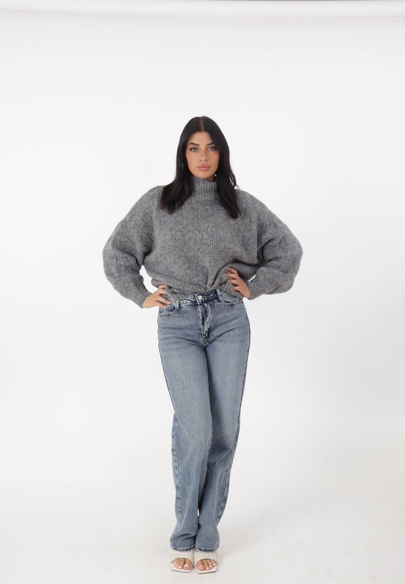 Grijze, oversized gebreide trui met een hoge halslijn, gecombineerd met lichtblauwe, high-waisted jeans. Model draagt lichtgekleurde sandalen.