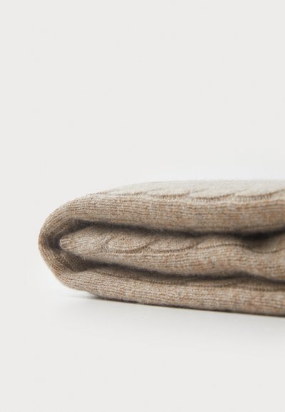 Beige virkkaustekniikalla valmistettu peitto, pehmeä pinta, taiteltuna esille pantuna, jossa on kaapelinvyyhti-kuviointi ja sävyjen hienovaraisia vaihteluita.