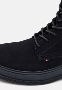 Botín de ante negro con punta redonda, cordones grises y suela de goma texturizada. Presenta un pequeño logotipo de color en el lateral.