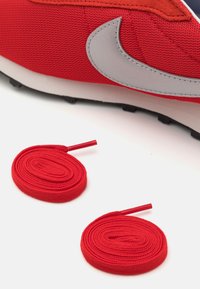 Crvena Nike tenisica s mrežastim gornjim dijelom i siva akcenta, uz dvije crvene okrugle vezice koje su smještene pored na bijeloj pozadini.