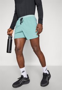 Lichtblauwe sportshorts met zwarte trekkoord en Nike-logo. Draag met zwarte sneakers, witte Nike-sokken en een zwarte waterfles in de hand.