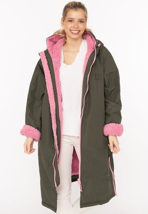 Young woman smiling, wearing a long dark green coat with pink fleece lining and cuffs, standing against a plain white background.