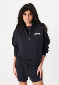 Crna dukserica s polu-zip ovratnikom i bijelim "ELLESSE" logotipom. Rebrasti manžeti, uparen s crnim kratkim hlačama koje imaju elastičnu struk.