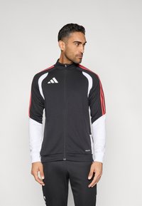 Fekete sportdzseki piros csíkokkal és fehér díszítésekkel, elülső cipzárral és álló gallérral. Simább, könnyű anyagból készült.