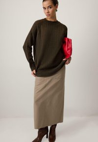 Adīta brūna džemperis ar ribotu tekstūru, saskaņota ar bēša midi svārkiem. Modelis tur sarkanu somiņu; valkā brūnas augstpapēžu kurpes.